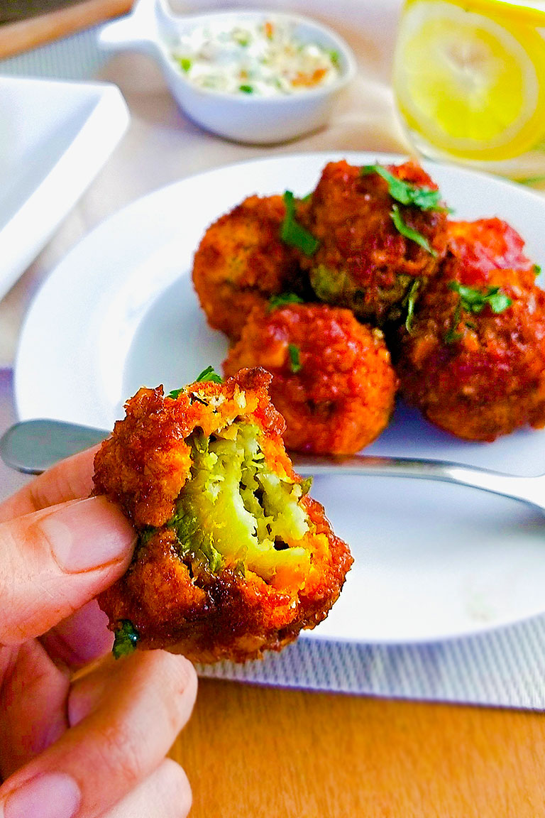 Close-up of a hand holding a fried chili brussels sprouts with a bite taken out, revealing green inner. Plate of more balls, dip, skillet, and lemon drink nearby.