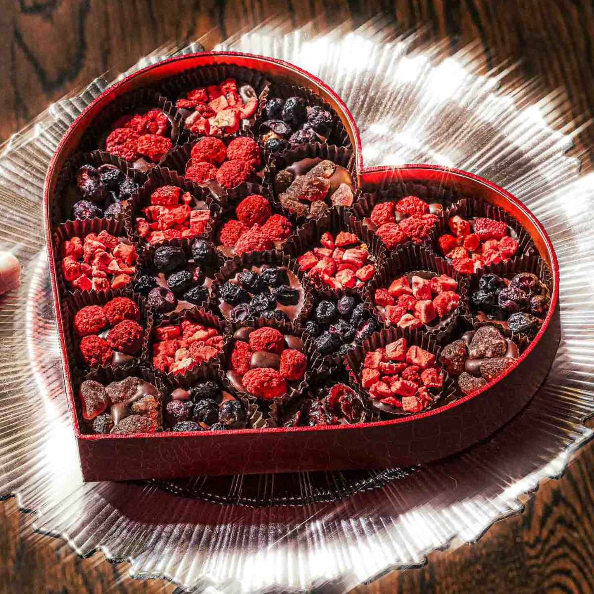 A heart-shaped box filled with assorted chocolates topped with dried berries, displayed on a clear glass plate over a wooden surface.
