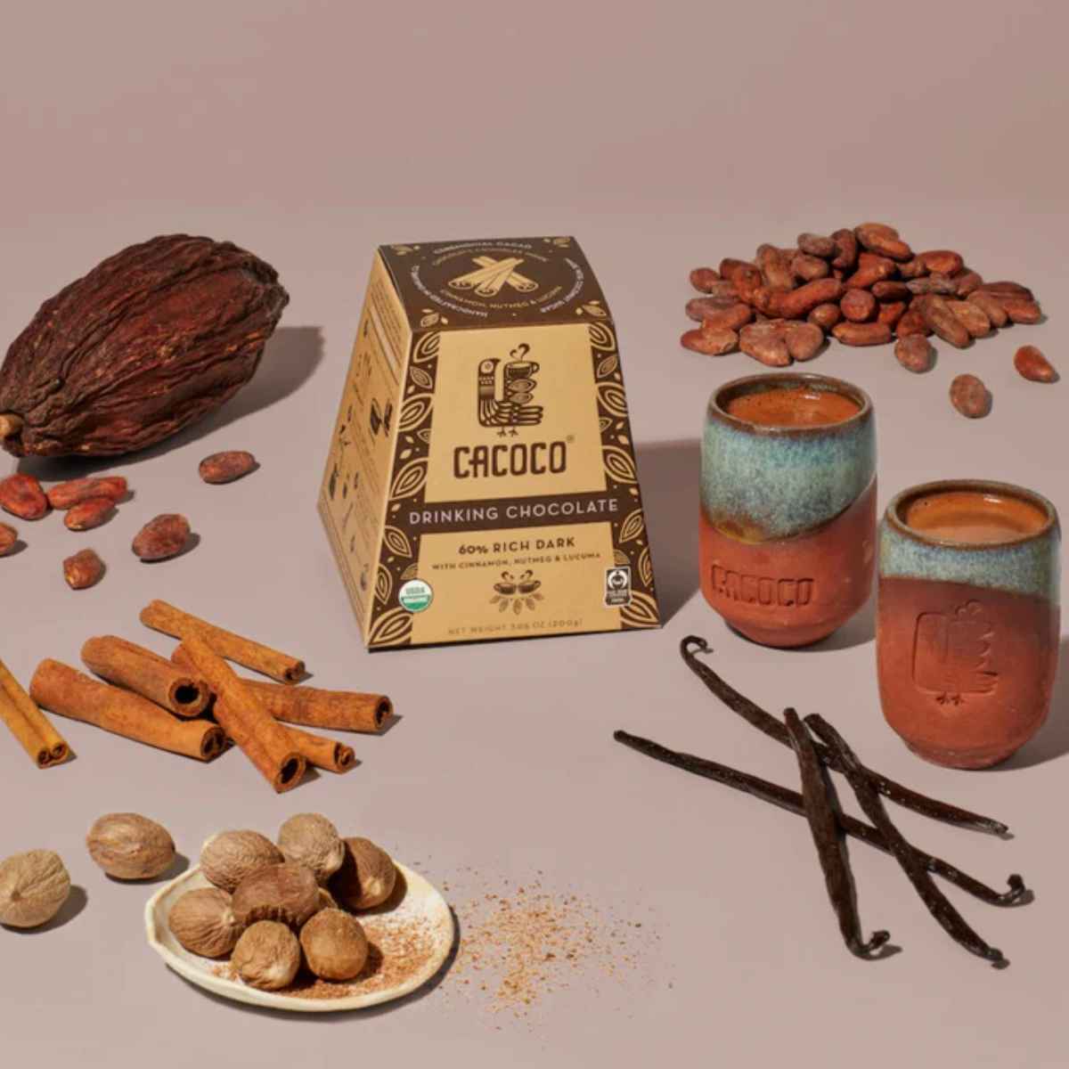Cacoco drinking chocolate box with two cups, cacao pods, cocoa beans, cinnamon sticks, nutmeg, and vanilla pods arranged on a neutral background.