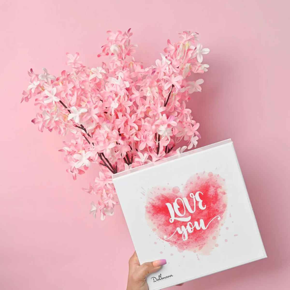 A hand holding a photo vegan chocolate box with "LOVE YOU" on its cover, surrounded by pink synthetic flowers against a pink backdrop.