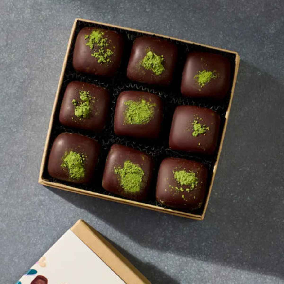 A box of nine chocolate truffles topped with green matcha powder, resting on a dark surface.