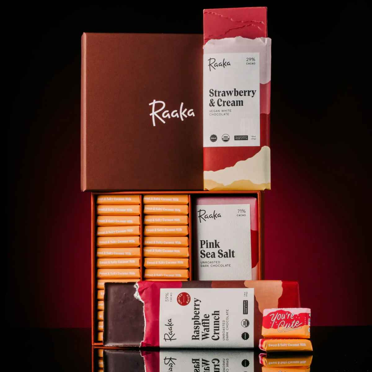 Raaka chocolate bars—Strawberry & Cream, Pink Sea Salt, and Raspberry Waffle Crunch—are arranged in and around an open brown gift box on a reflective surface.