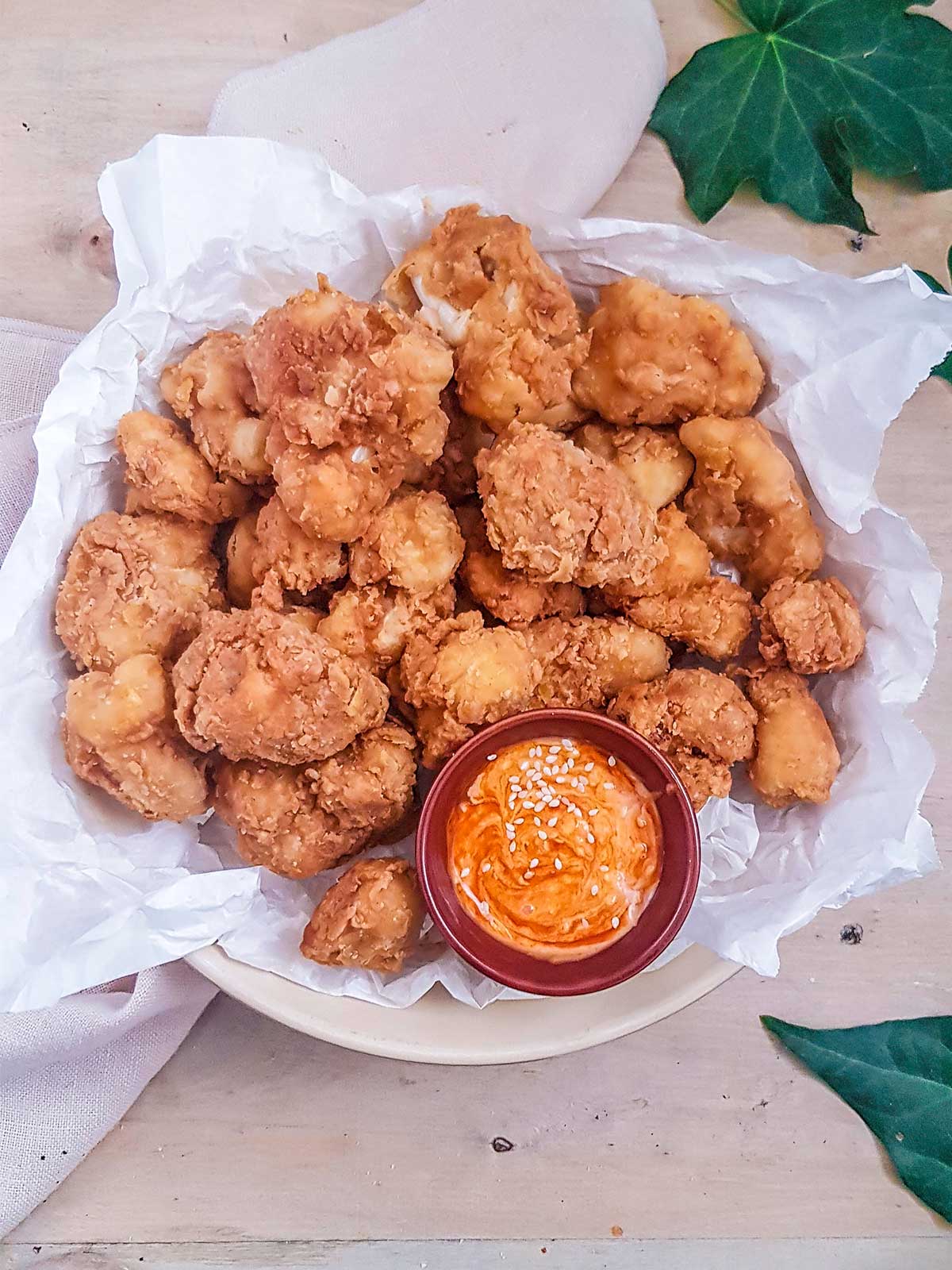 A plate of crispy fried cauliflower bites rests on crumpled white paper, beside a small red dish of creamy orange sauce with sesame seeds, evoking a casual, appetizing vibe.