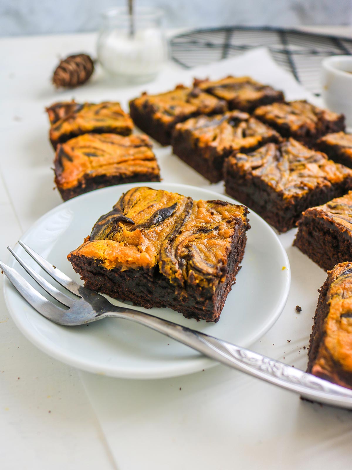 Chocolate brownies with a marbled top of swirled pmpkiin are arranged on a white plate and table. A fork is placed beside the plate.