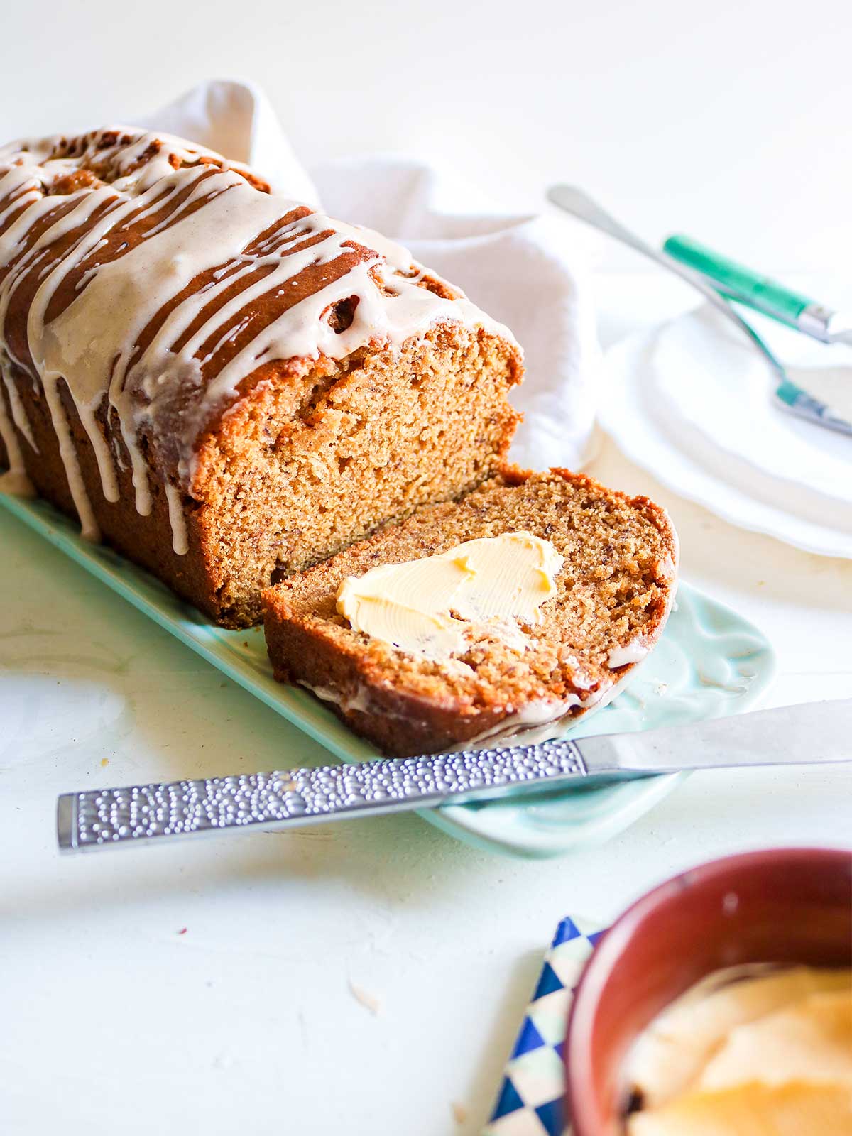 A loaf of iced, sliced pumpkin banana bread on a teal platter with a dollop of butter on one slice. A knife beside it; warm and inviting atmosphere.