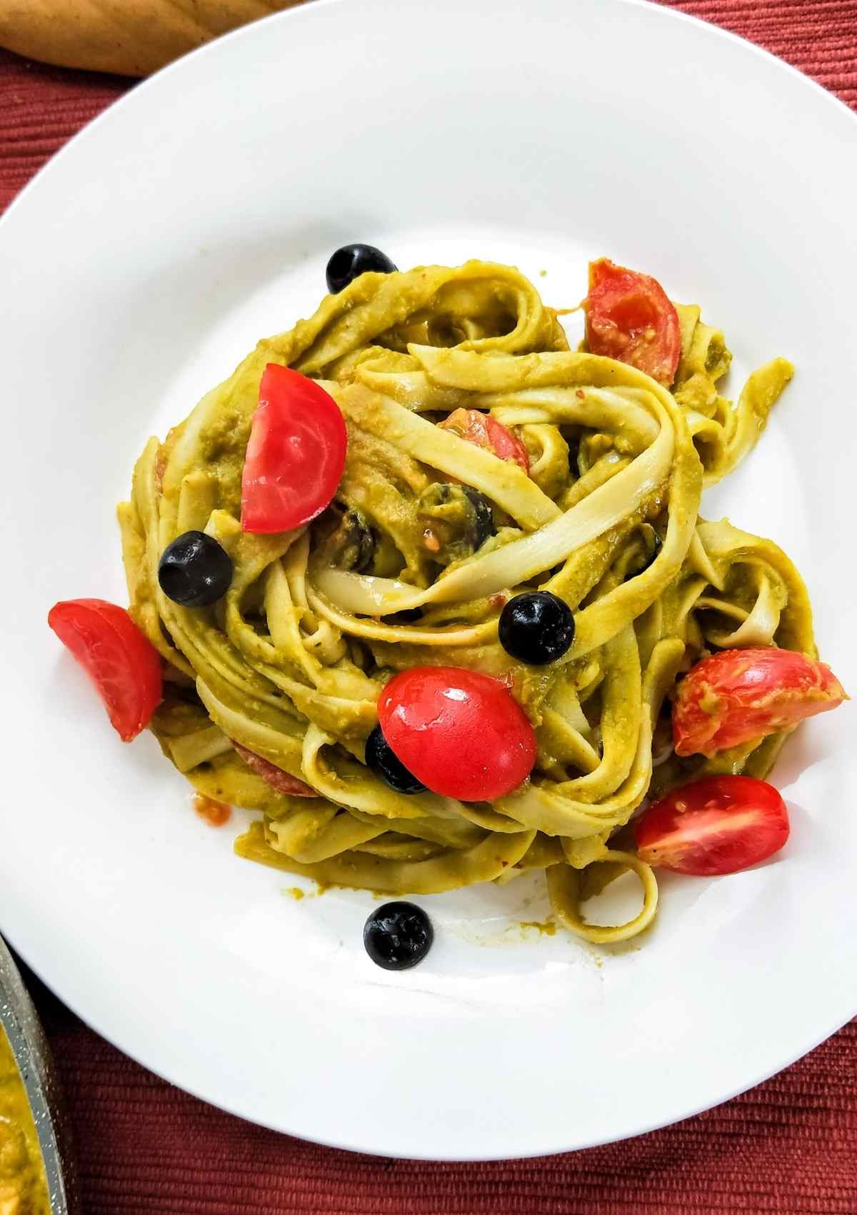 vegan spinach pasta served in a white plate topped with black olives and red cherry tomatoes