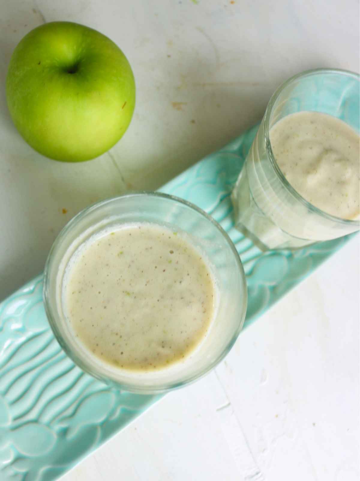Green apple smoothie served in two glasses placed on a tray and green apple on the background.