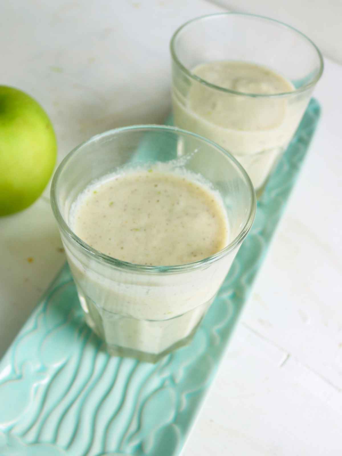 Green apple smoothie served in two glasses placed on a tray and green apple on the background.