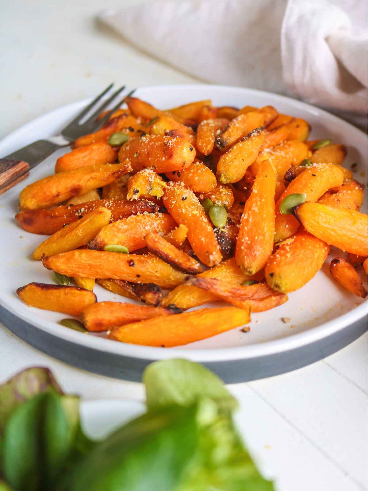 Orange color baby carrots served in a white plate topped with pumpkin seeds and vegan parmesan.