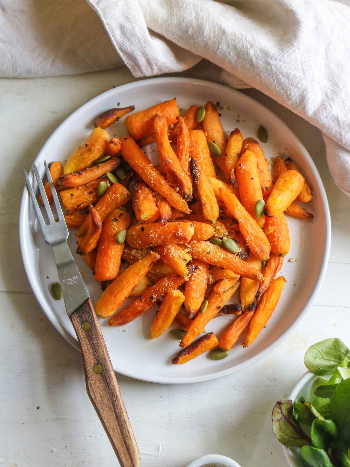 Orange color baby carrots served in a white plate and a white napkin in background and wooden fork on side.