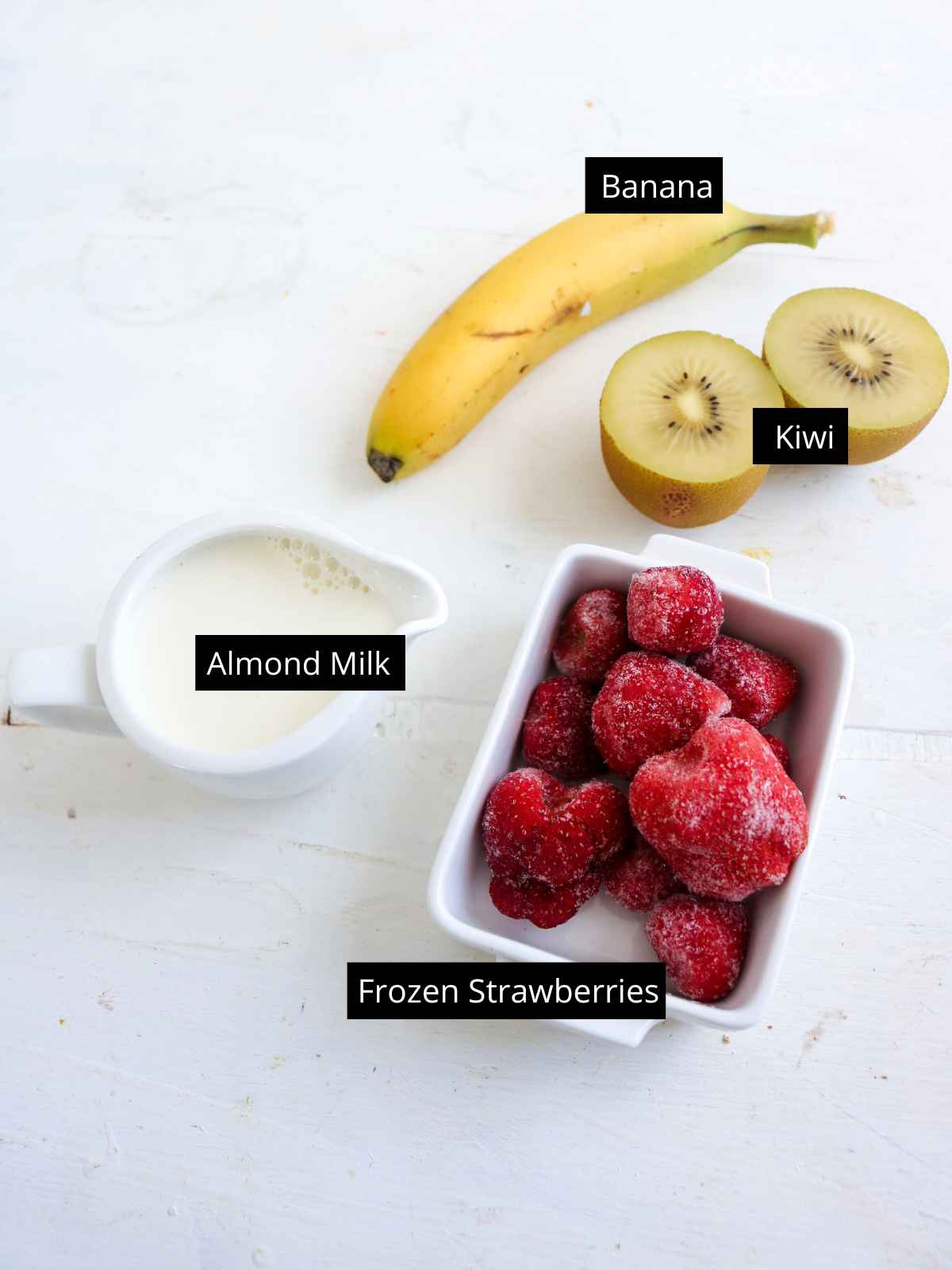 yellow banana, 2 kiwi halves, almond milk in a white pot and frozen strawberries in a white dish placed on a white background.