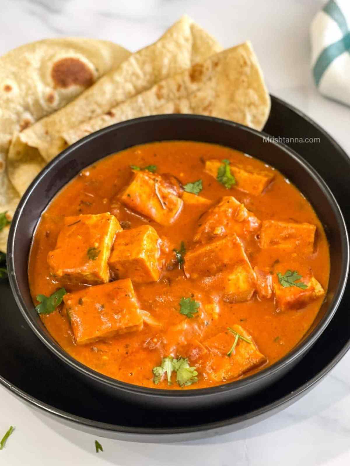 Brigh orange color tofu tikka masala served in a black color bowl. Naan bread on background.