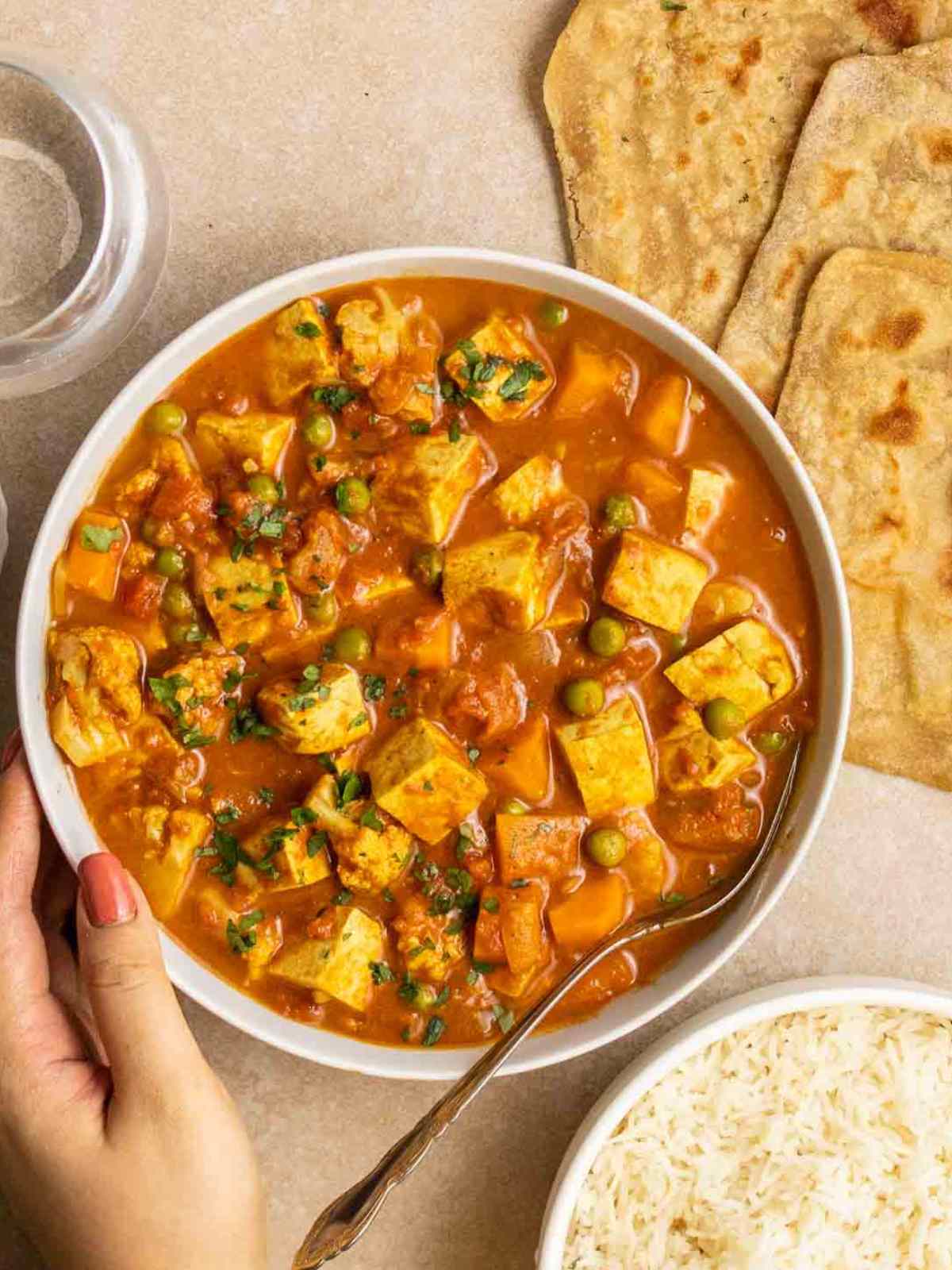 A hand is holding a bowl filled with tofu madras curry.