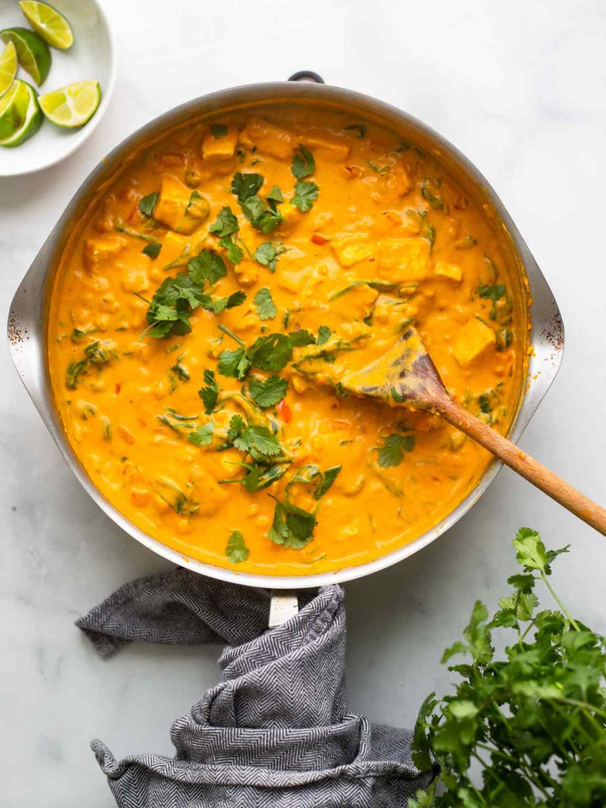 Bright orange color pumpkin tofu curry served in a metal pan with wooden spoon on side.