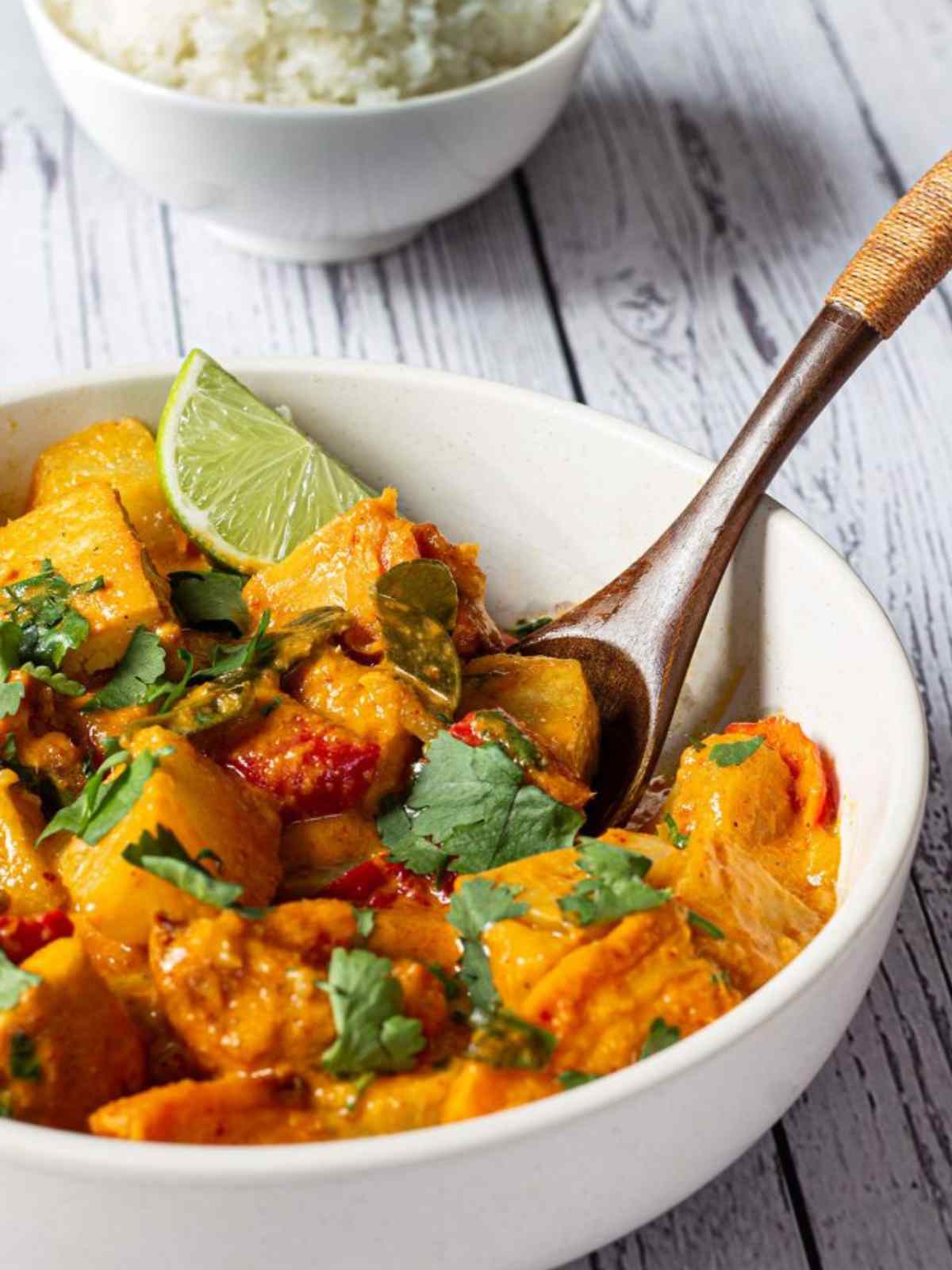 tofu and sweet potato curry served in a white bowl. A lemon slice and wooden spoon placed on side.