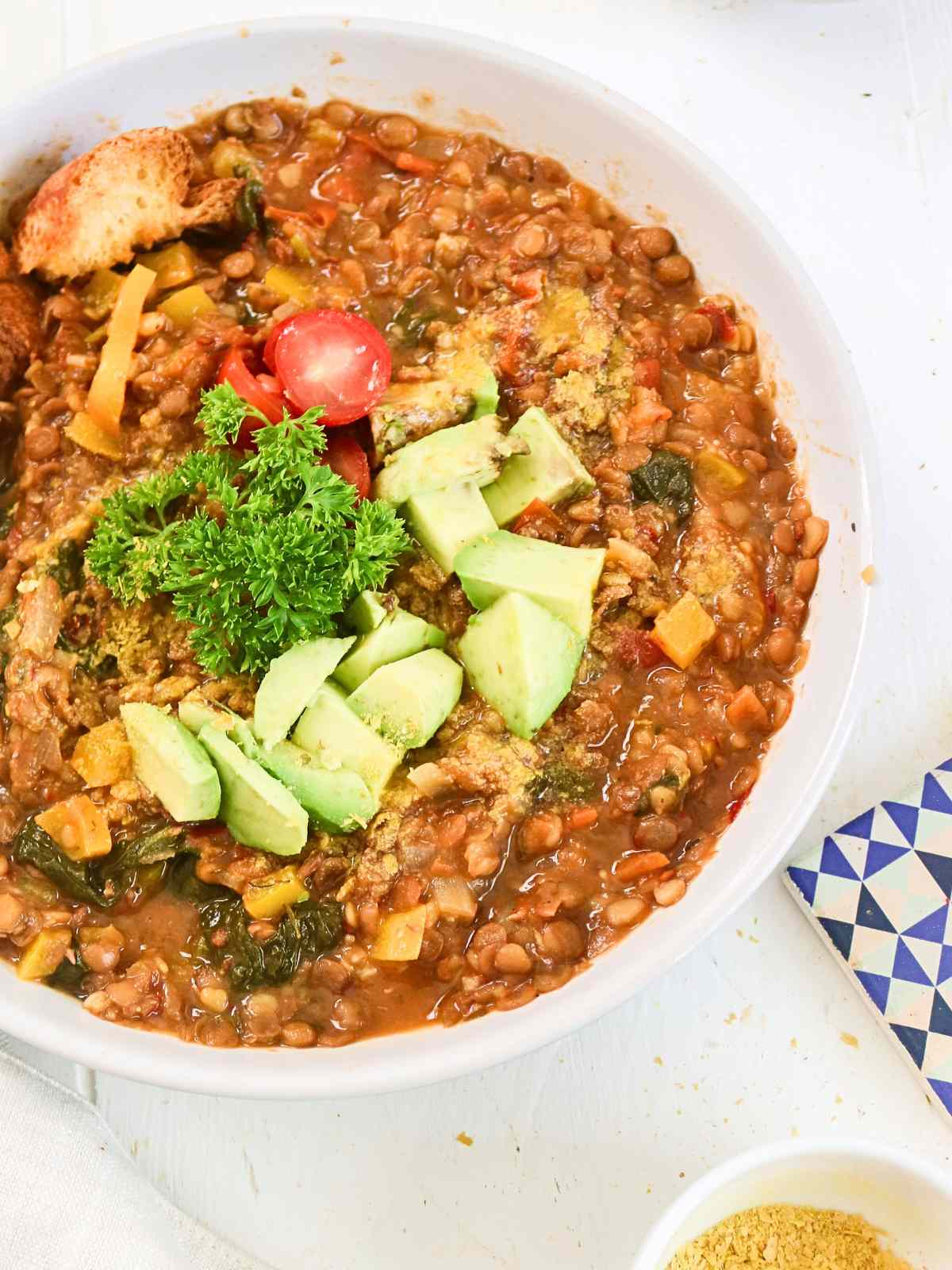 Lentil stew served in a bowl topped with avocado, parsley and cherry tomatoes.