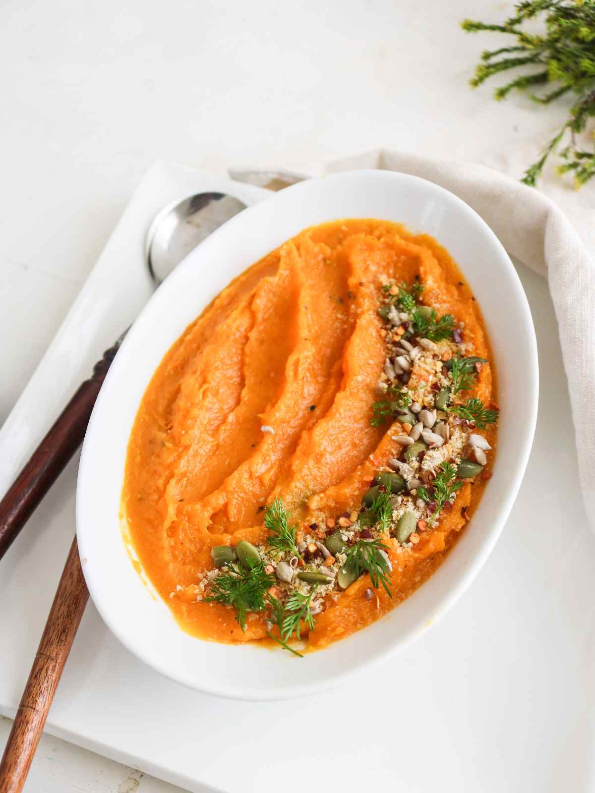 Orange color butternut squash mash served in a white dish garnish with fresh green herbs and seeds.