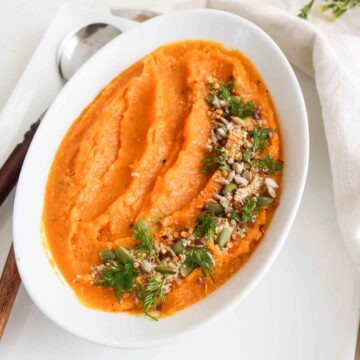 Orange color butternut squash mash served in a white dish garnish with fresh green herbs and seeds.