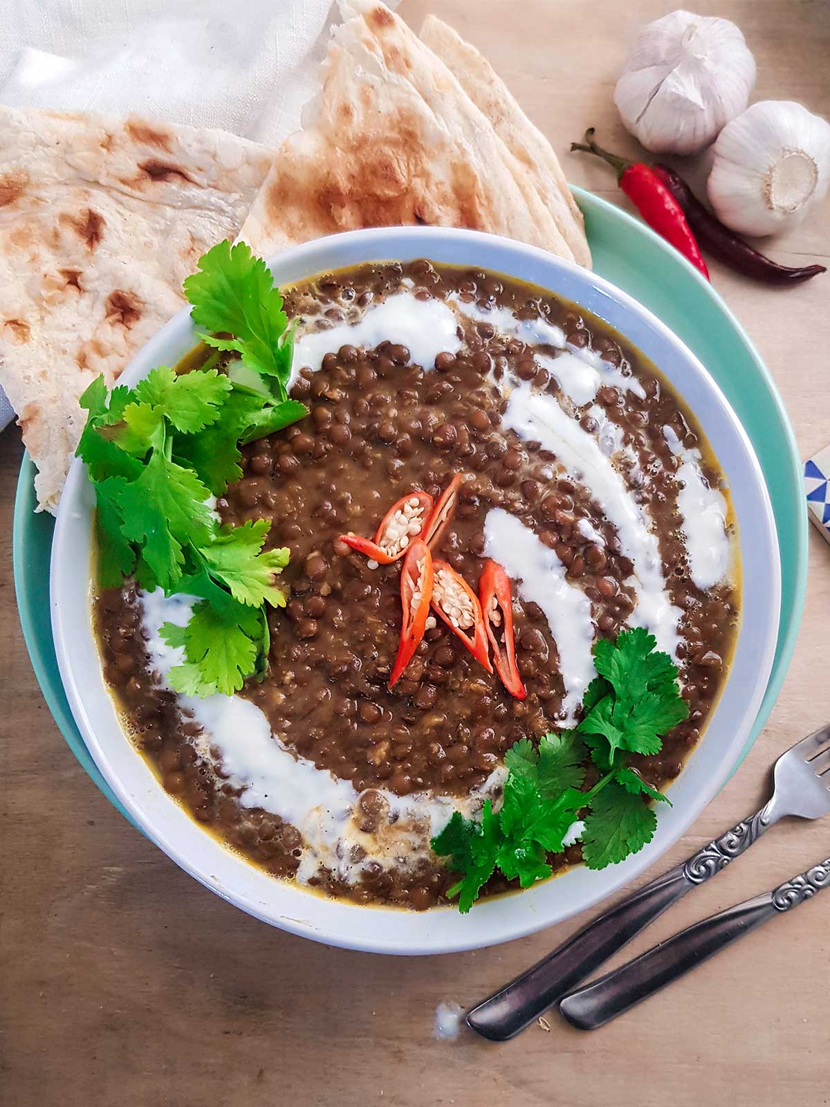 Brown lentil dahl served in a white bowl garnished with green cilantro leaves, coconut cream and red chilies.