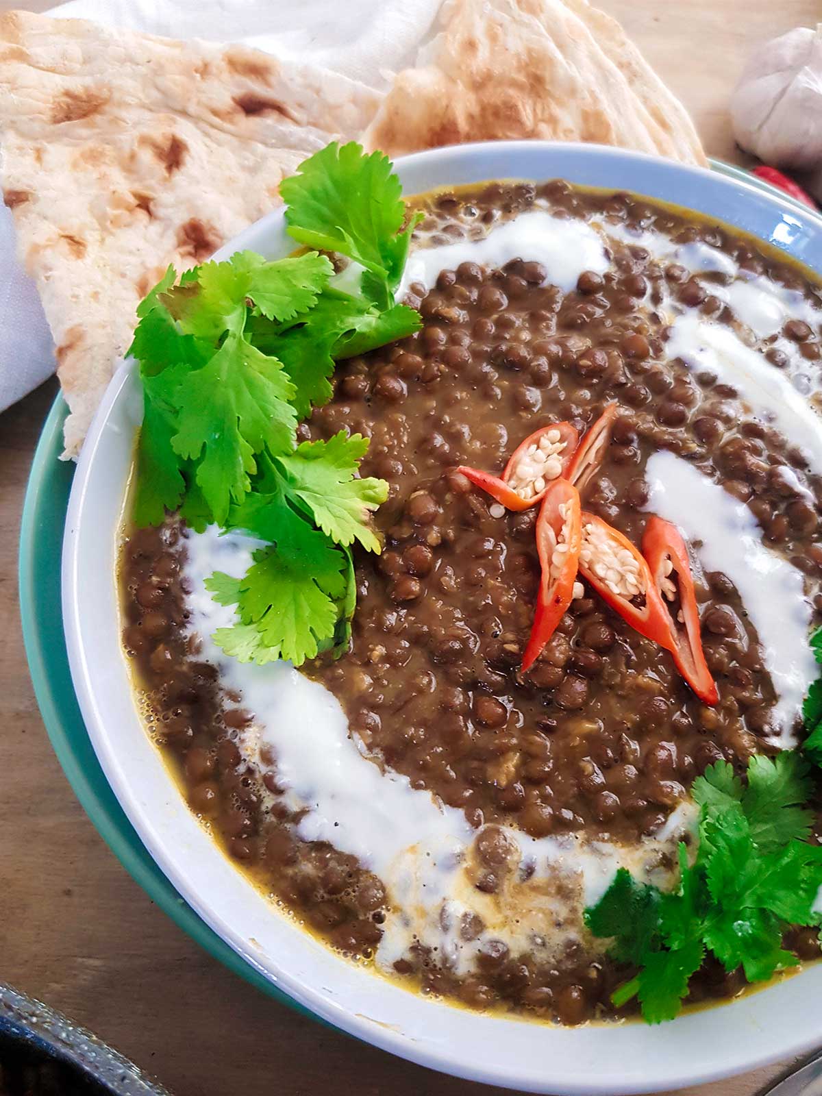 Whole masoor dal served in a white bowl garnished with green cilantro leaves, coconut cream and red chilies.