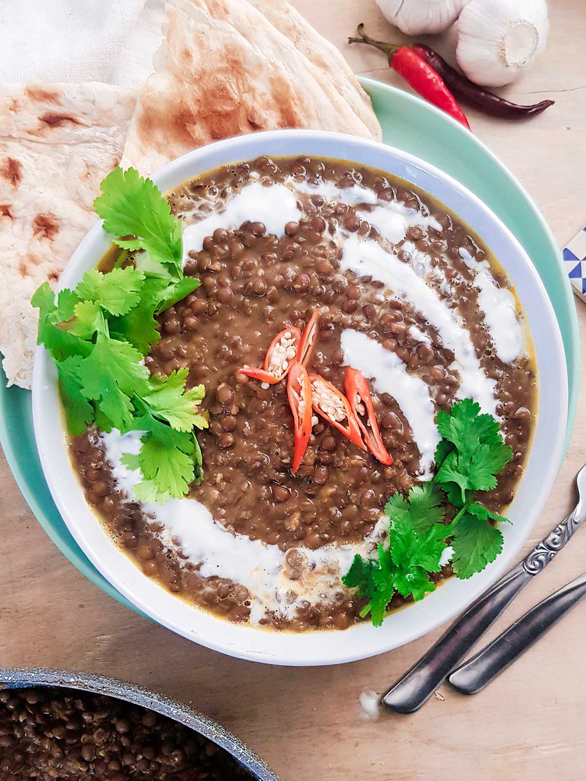 Brown lentil curry served in a white bowl garnished with green cilantro leaves, coconut cream and red chilies.