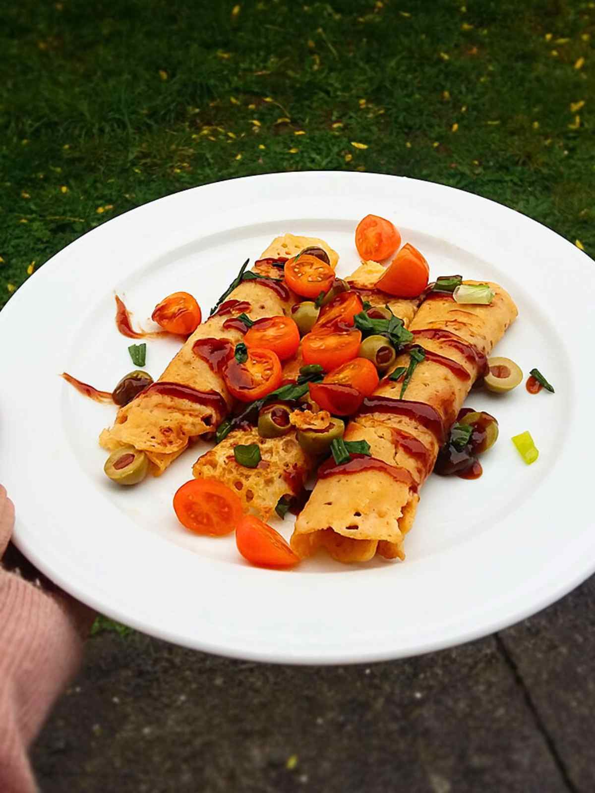 Holding  a plate of crepes topped with bbq sauce , cherry tomatoes and olives.