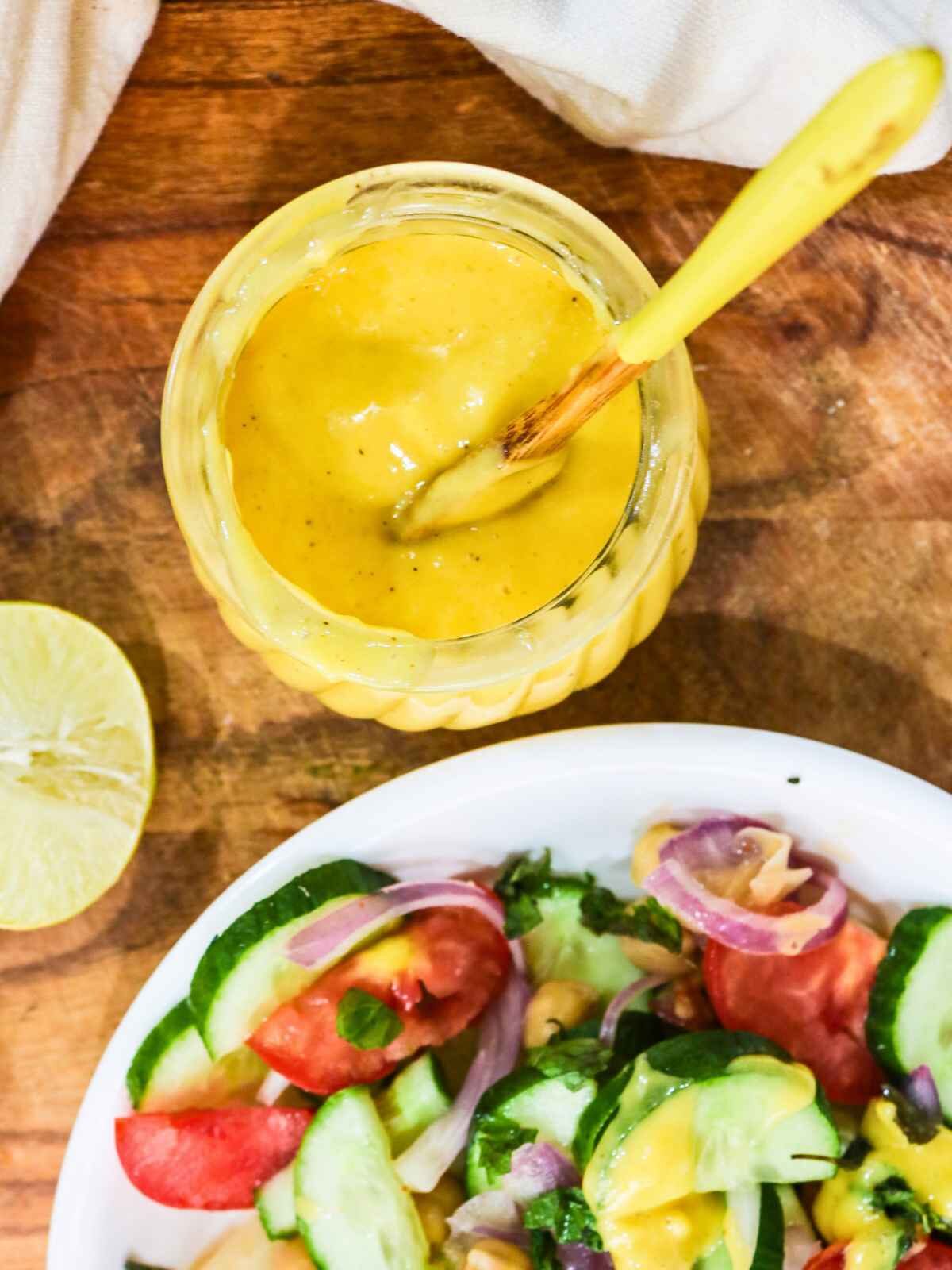 A glass jar of creamy yellow dressing with a spoon on a wooden board beside a fresh salad with cucumbers, tomatoes, red onions, and half a lime.