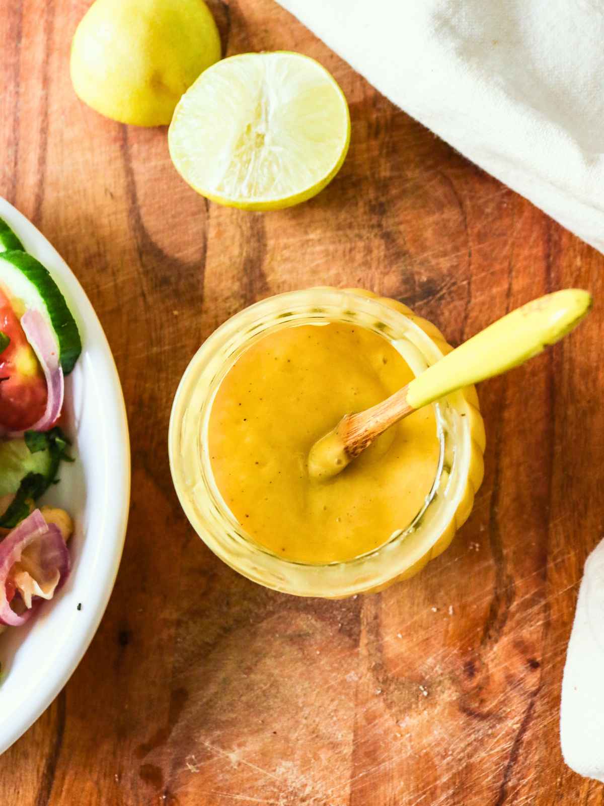A jar of creamy yellow dressing with a spoon, on a wooden surface, next to a halved lemon and salad, conveying a fresh, homemade vibe.