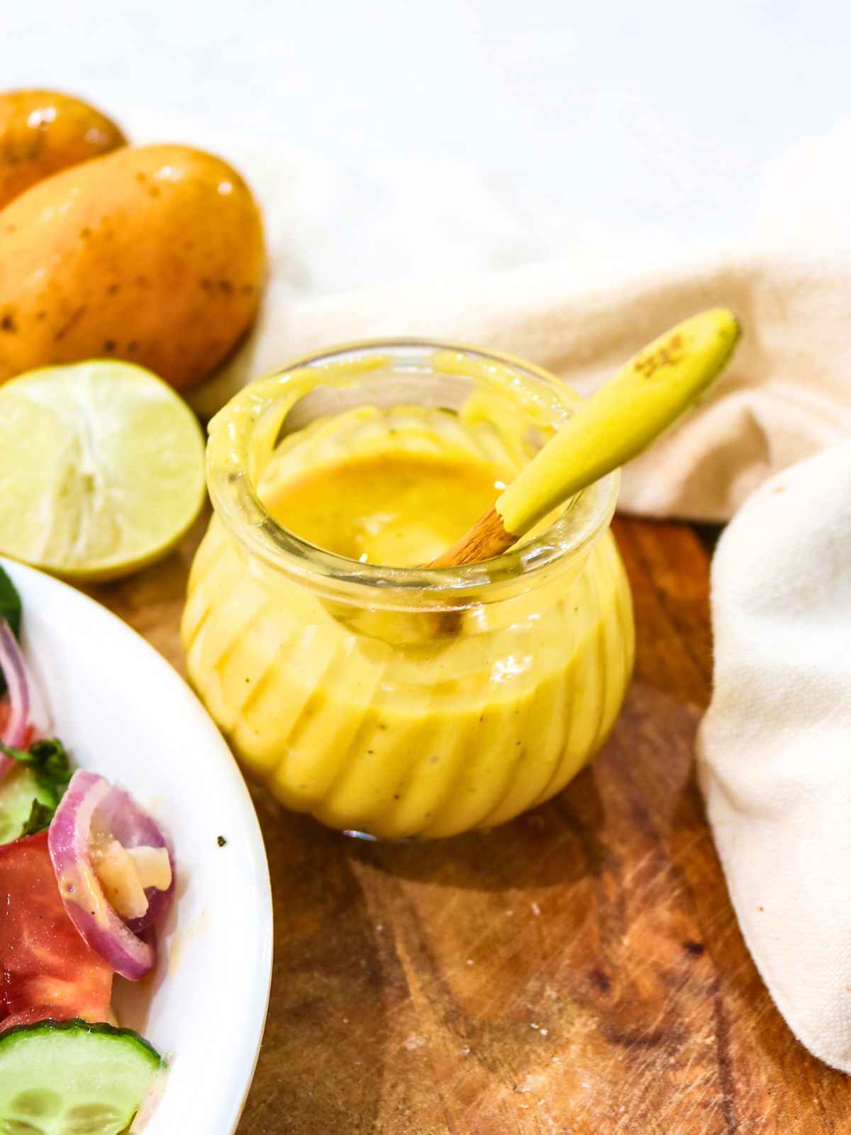 A jar of vibrant yellow mango salad dressing on a wooden surface with a spoon inside. Fresh salad and fruit, including cucumber, tomato, and mango, surround it.