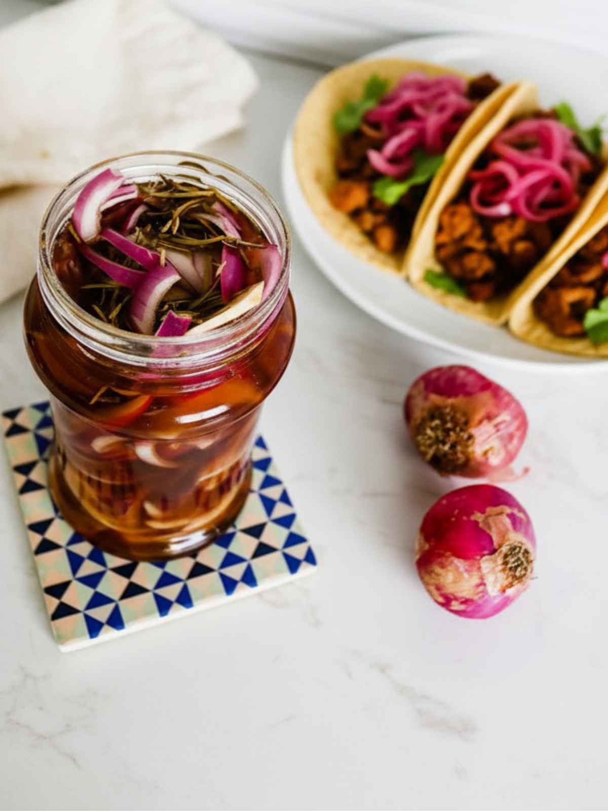 Jar of pickled onions with rosemary on a geometric coaster, accompanied by fresh radishes. In the background, tacos topped with balsamic pickled onions.