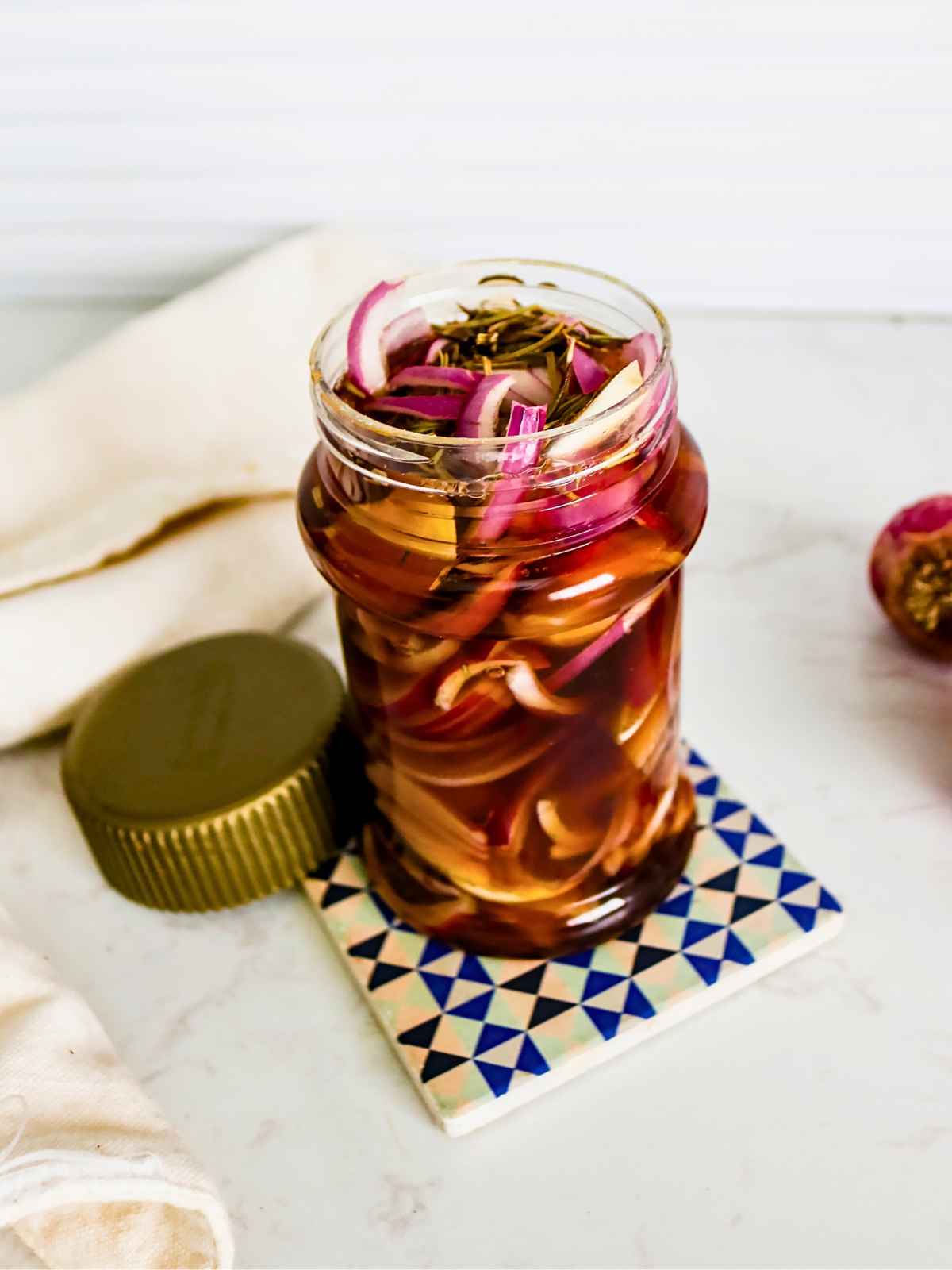 A jar of pickled red onions is open on a geometric-patterned coaster. The golden lid is placed beside it, and a white cloth partially drapes underneath.