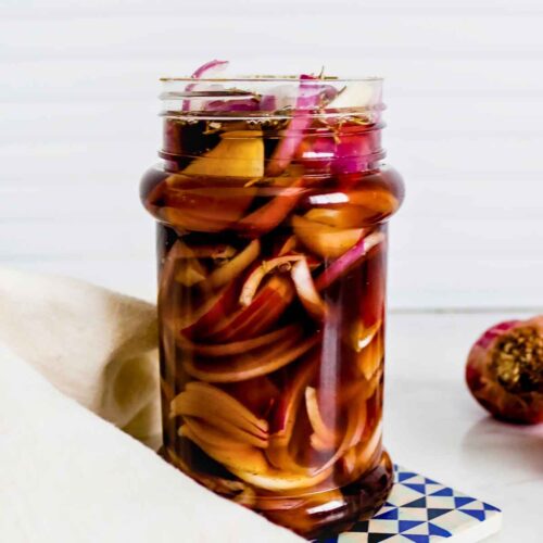 A glass jar filled with pickled red onions in vinegar brine. The jar is set on a blue and white coaster, with a light napkin nearby and a sliced onion in the background.