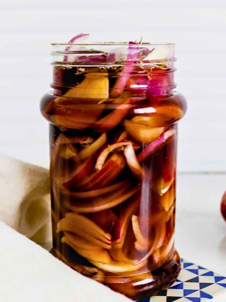 A glass jar filled with pickled red onions in vinegar brine. The jar is set on a blue and white coaster, with a light napkin nearby and a sliced onion in the background.