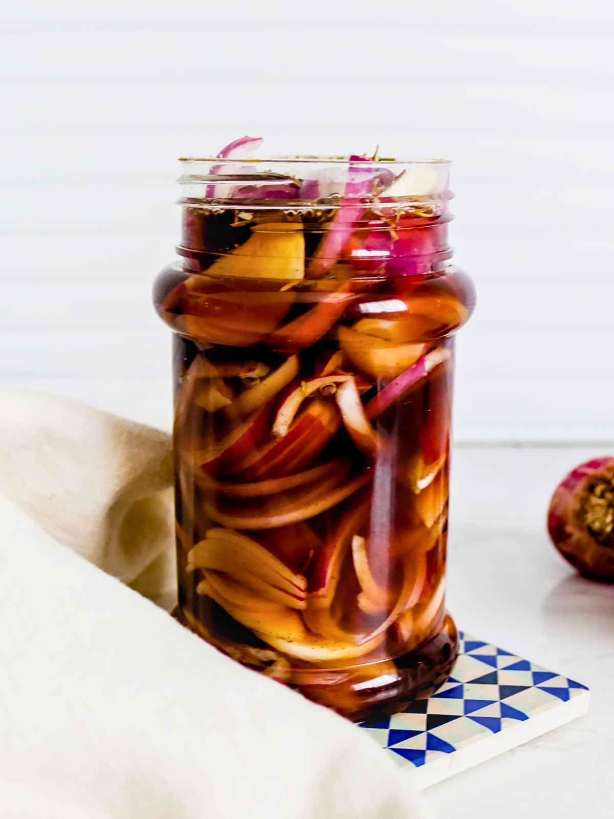 A glass jar filled with pickled red onions in vinegar brine. The jar is set on a blue and white coaster, with a light napkin nearby and a sliced onion in the background.